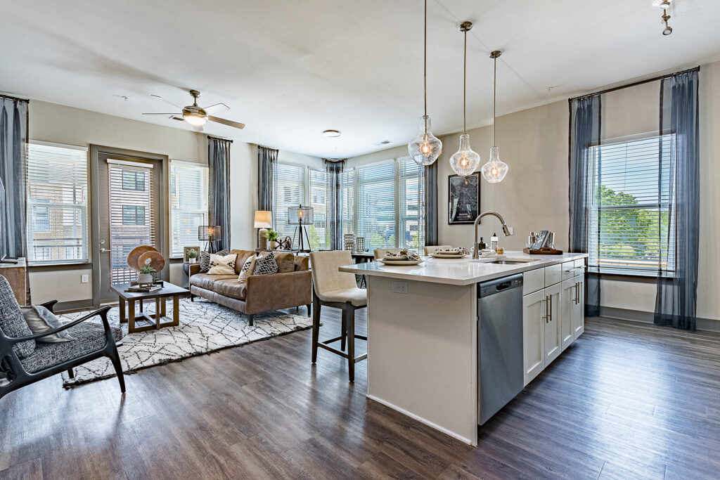 A modern living room and kitchen with a gray sofa, brown armchair, wooden coffee table, and gray island with pendant lights.