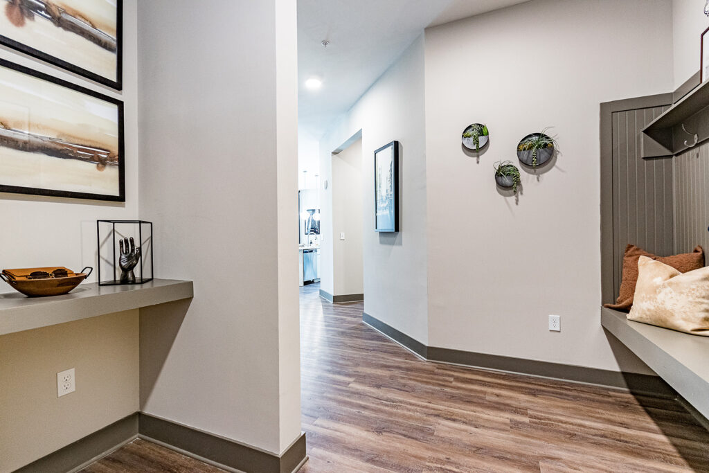 A modern hallway with light gray walls, dark wood flooring, framed pictures, a bench with a pillow, and a shelf with plants.