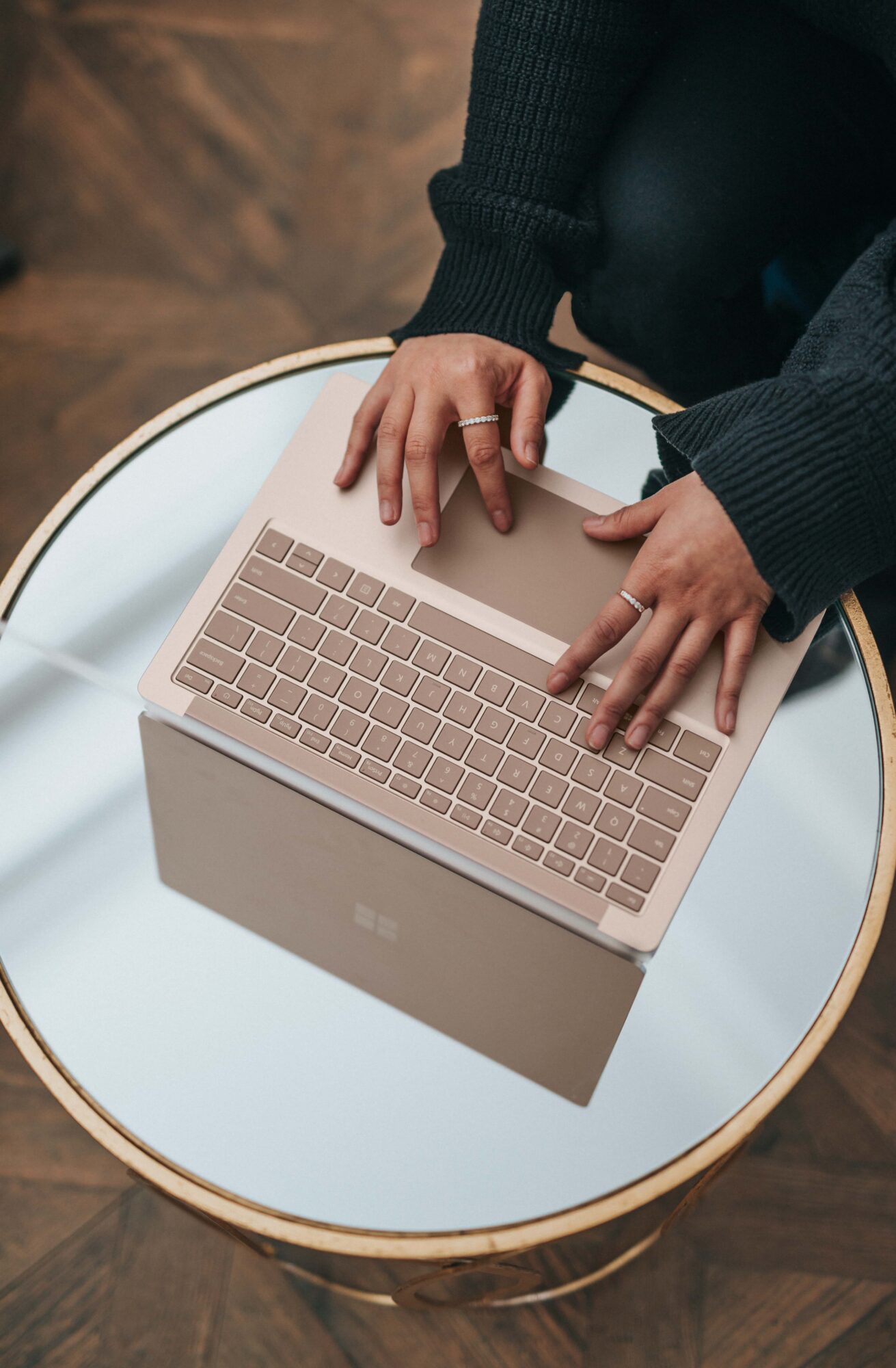 A person’s hands typing on a rose gold laptop on a round white table with a gold rim, with a wooden floor in the background.