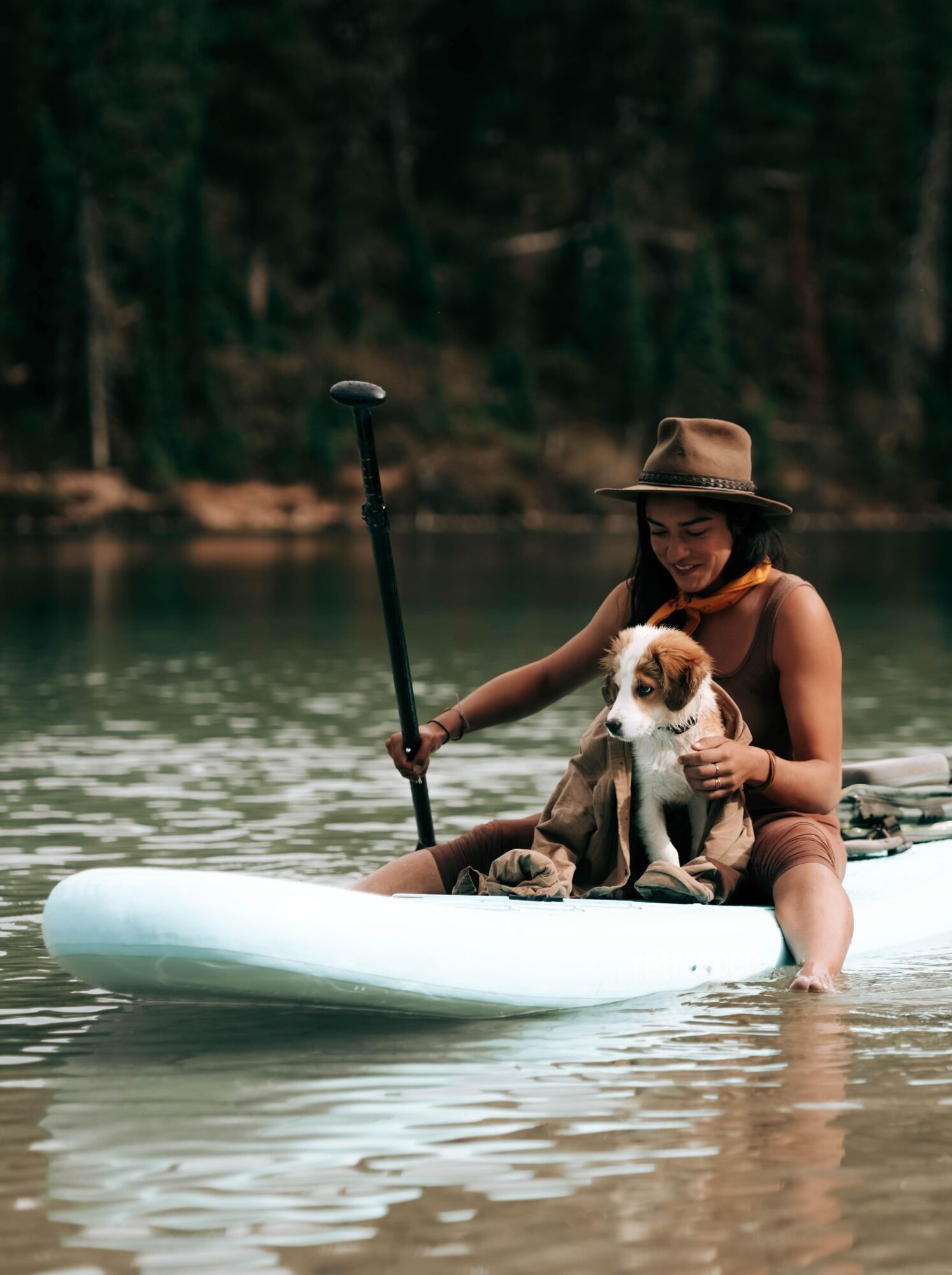 A person and a dog on a paddle board in a calm lake, surrounded by trees and a shoreline.