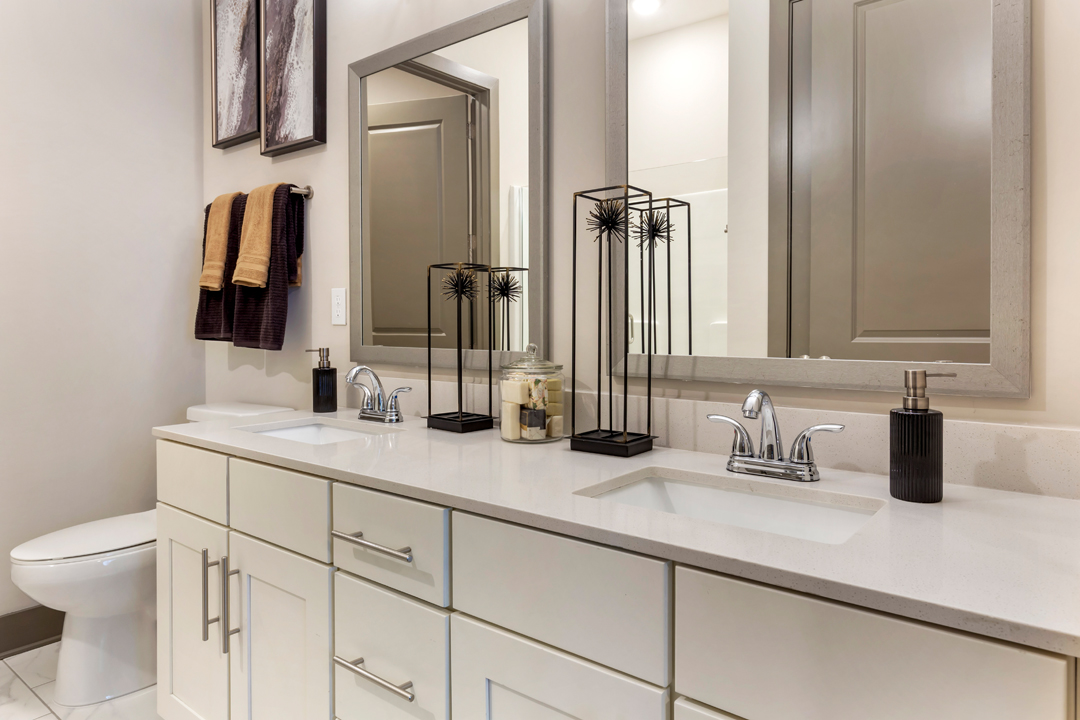 Modern bathroom with white countertop, two sinks, mirror, black candle holders, and soap dispenser.